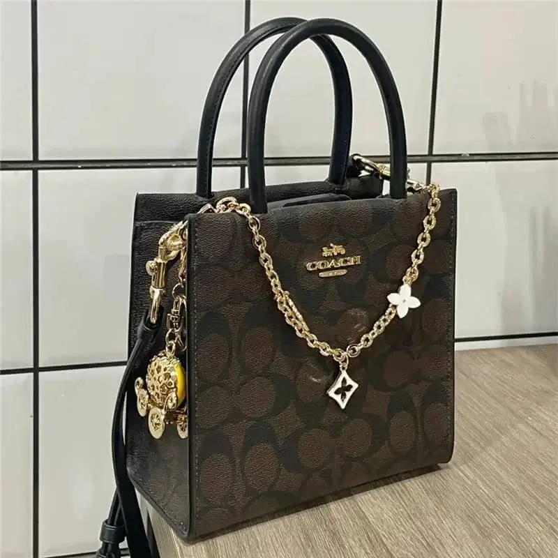 A classic COACH tote bag featuring a dark brown signature monogram leather, adorned with a gold chain detail and elegant charms including floral and diamond-shaped pendants, combining timeless elegance with modern style for everyday wear or casual outings.